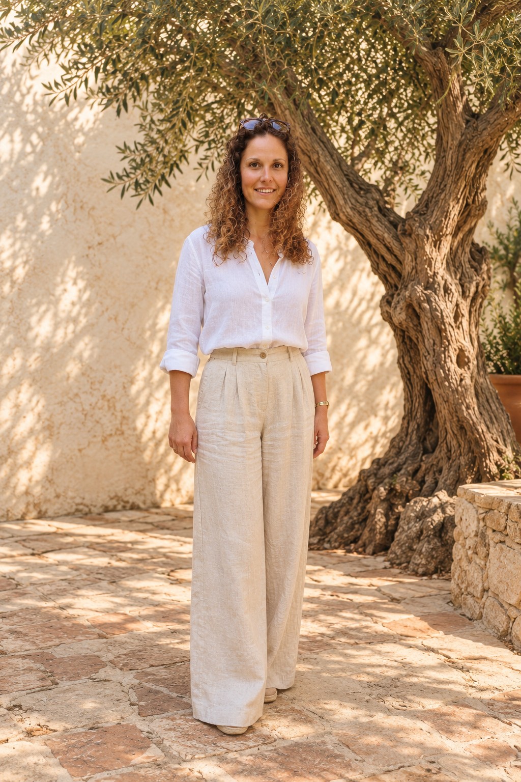 Cassie, founder of Rise & Root, in a white linen shirt and beige trousers beside a large olive tree